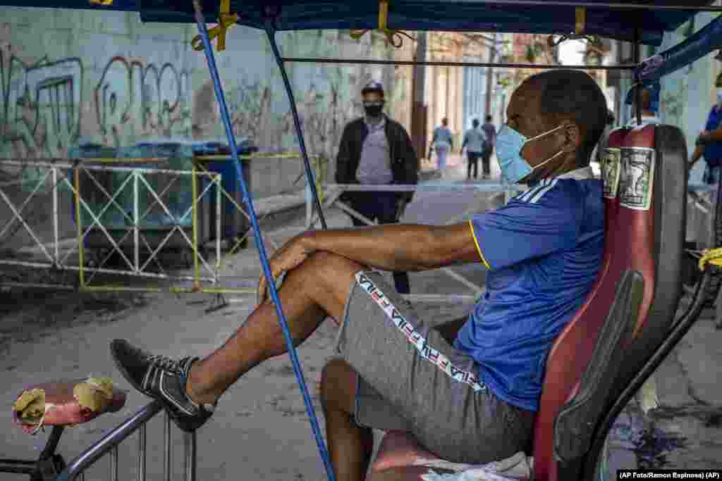 Un taxista en bicicleta espera clientes afuera de una puerta donde la policía controla el acceso a un vecindario como una forma de frenar la propagación de COVID-19 en La Habana, Cuba, el lunes 22 de febrero de 2021. (AP Foto/Ramon Espinosa)