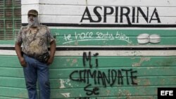 Un hombre con la cara tapada descansa junto a una pared con una pintada en la que se lee "Mi comandante se queda".