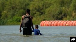 Migrantes caminan junto a grandes boyas que se utilizan como una barrera fronteriza flotante en el Río Grande, en Eagle Pass, Texas. (Foto AP/Eric Gay)